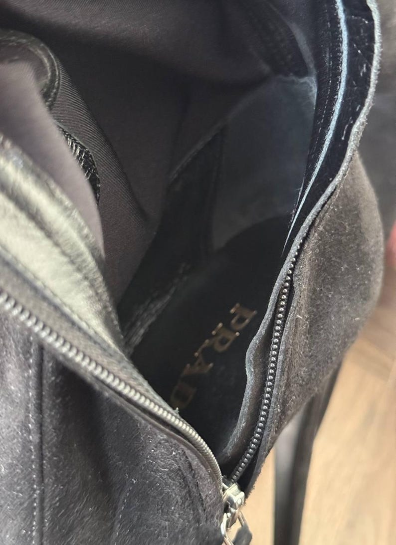 May include: Close-up of a black suede boot interior with a zipper detail. The brand name "PRADA" is visible on the insole. The boot has a high heel and a sleek, modern design.