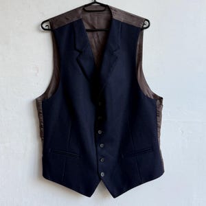 Può includere: Un gilet blu navy con schiena e revers in raso marrone, appeso a una gruccia nera. Il gilet presenta un design classico con colletto a lancia, chiusura con bottoni e due tasche anteriori. È un capo formale.