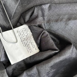 Puede incluir: Primer plano de tela gris con brillo met&aacute;lico. Una etiqueta blanca est&aacute; sujeta con un clip de metal plateado. La etiqueta tiene texto japon&eacute;s. La tela est&aacute; doblada y tiene costuras visibles.