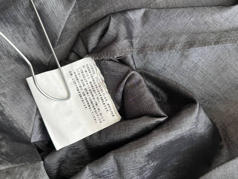 Puede incluir: Primer plano de tela gris con brillo met&aacute;lico. Una etiqueta blanca est&aacute; sujeta con un clip de metal plateado. La etiqueta tiene texto japon&eacute;s. La tela est&aacute; doblada y tiene costuras visibles.