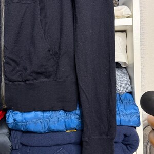 May include: A black zip-up hoodie with a ribbed hem and cuffs. The hoodie has a kangaroo pocket and a silver zipper. The sleeve is visible, and the garment is folded on a shelf with other clothing.