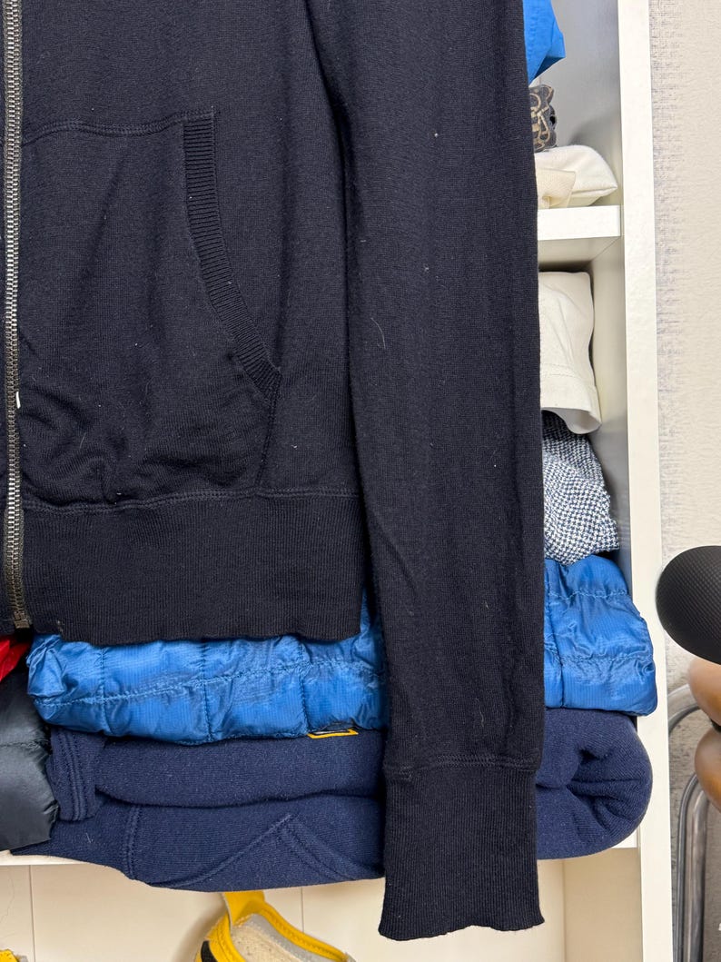 May include: A black zip-up hoodie with a ribbed hem and cuffs. The hoodie has a kangaroo pocket and a silver zipper. The sleeve is visible, and the garment is folded on a shelf with other clothing.
