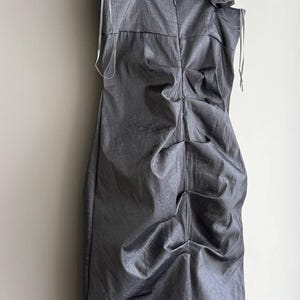 Puede incluir: Un vestido gris sin mangas con cuello en V y un dise&ntilde;o fruncido y drapeado. El vestido tiene un detalle de volantes &uacute;nico en un hombro y est&aacute; colgado de una percha de madera. La tela tiene un brillo sutil.