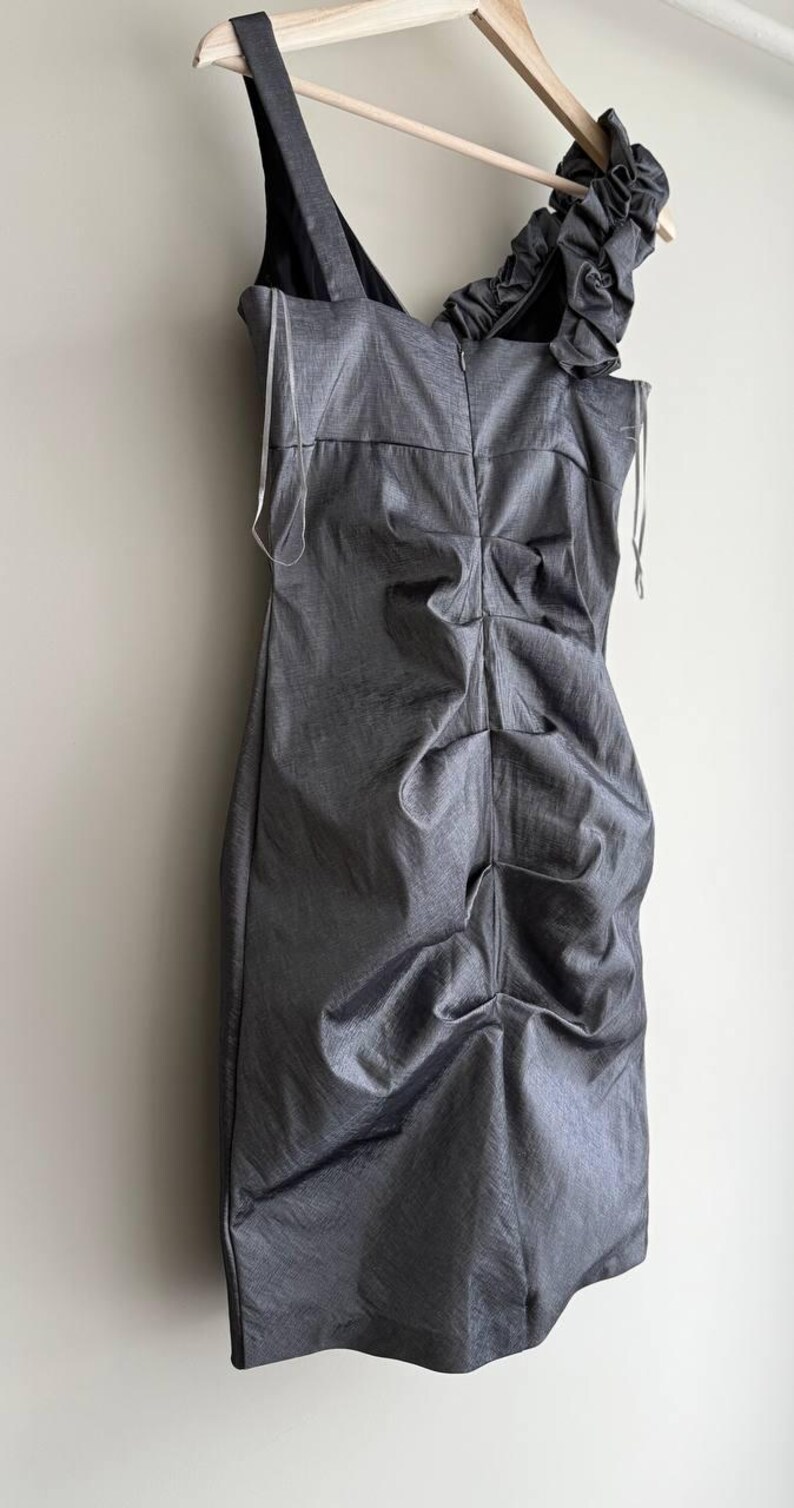 Puede incluir: Un vestido gris sin mangas con cuello en V y un dise&ntilde;o fruncido y drapeado. El vestido tiene un detalle de volantes &uacute;nico en un hombro y est&aacute; colgado de una percha de madera. La tela tiene un brillo sutil.