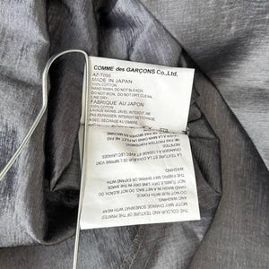 Puede incluir: Primer plano de una prenda gris con una etiqueta de Comme des Gar&ccedil;ons Co., Ltd. La etiqueta incluye instrucciones de lavado y el texto "MADE IN JAPAN". La tela tiene un brillo sutil y est&aacute; drapeada sobre una percha.