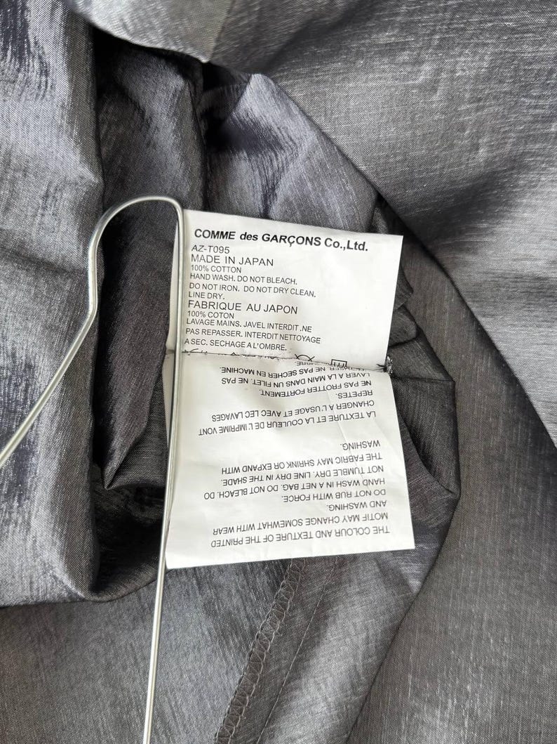 Puede incluir: Primer plano de una prenda gris con una etiqueta de Comme des Gar&ccedil;ons Co., Ltd. La etiqueta incluye instrucciones de lavado y el texto "MADE IN JAPAN". La tela tiene un brillo sutil y est&aacute; drapeada sobre una percha.