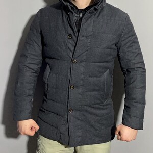 May include: A dark gray quilted jacket with a button-up front and a high collar. The jacket has a textured fabric and a slightly longer length.