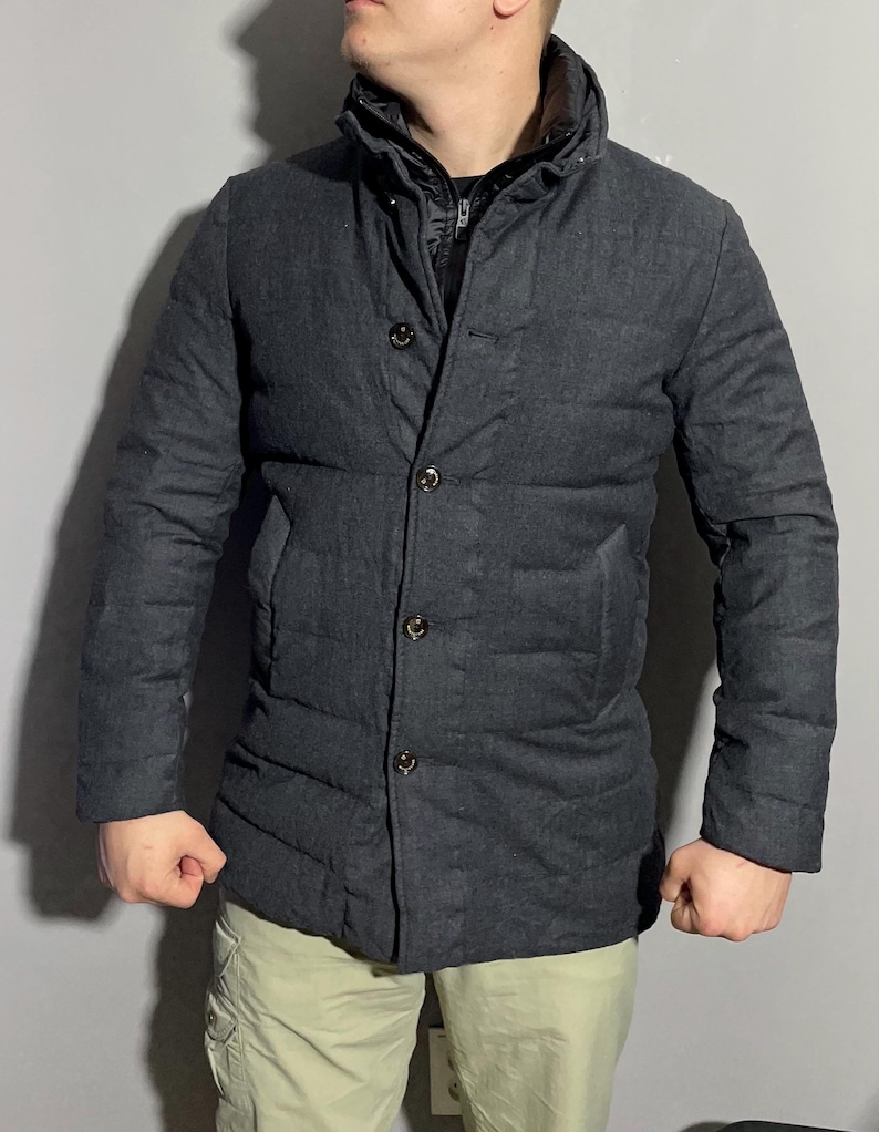 May include: A dark gray quilted jacket with a button-up front and a high collar. The jacket has a textured fabric and a slightly longer length.