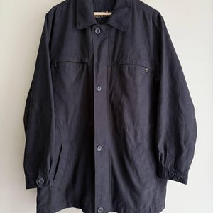 May include: A dark navy blue jacket with a collared neckline, button-down front, and multiple pockets. The jacket is hanging on a wooden hanger against a neutral background. The sleeves have buttoned cuffs.