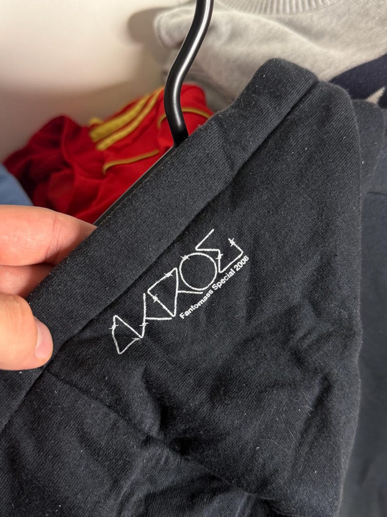 Pode incluir: T-shirt preto com texto bordado em branco. O texto diz "AKROS Fantomas Special 2008". A t-shirt est&aacute; pendurada num cabide preto. Outras pe&ccedil;as de roupa s&atilde;o vis&iacute;veis no fundo.