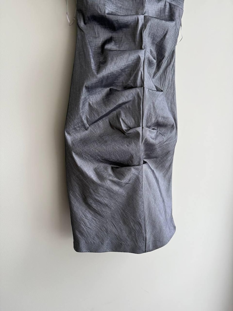 Puede incluir: Un vestido gris sin tirantes con un dise&ntilde;o fruncido. El vestido est&aacute; hecho de una tela brillante y texturizada y cuelga verticalmente contra una pared blanca. El vestido parece ser hasta la rodilla.