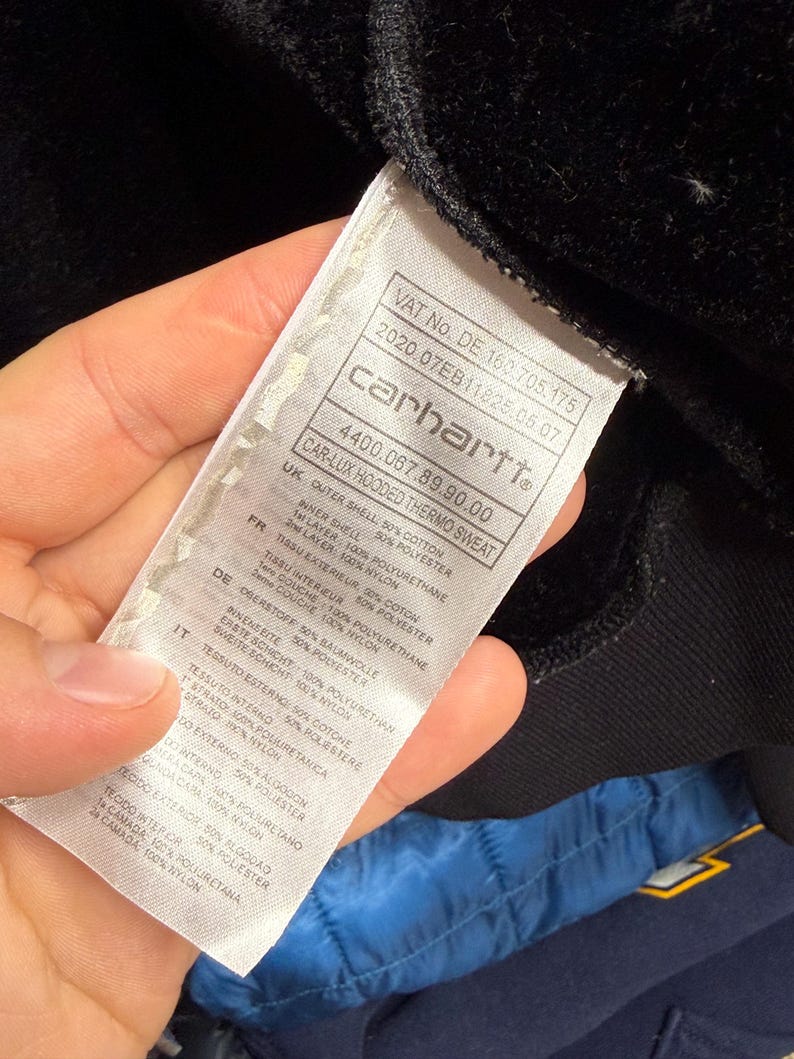 Pode incluir: Close-up de uma etiqueta Carhartt, com texto em v&aacute;rios idiomas. A etiqueta branca tem texto preto e inclui detalhes sobre os materiais e a composi&ccedil;&atilde;o da pe&ccedil;a de vestu&aacute;rio. A etiqueta &eacute; mantida contra um fundo de tecido preto.