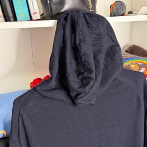 May include: A black hooded sweatshirt with a drawstring hood. The sweatshirt is made of a soft, knit fabric. The image is taken from the back, showing the hood and the top of the shoulders.
