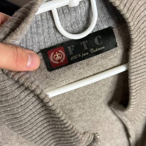 Pu&ograve; includere: Primo piano di un maglione in cashmere beige con colletto e polsini a coste. Il maglione &egrave; appeso a una gruccia bianca. &Egrave; visibile un'etichetta nera con la scritta "FTC 100% pure Cashmere".