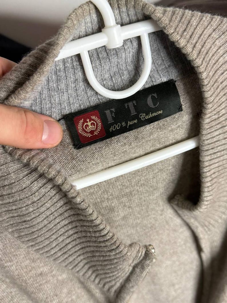 Pu&ograve; includere: Primo piano di un maglione in cashmere beige con colletto e polsini a coste. Il maglione &egrave; appeso a una gruccia bianca. &Egrave; visibile un'etichetta nera con la scritta "FTC 100% pure Cashmere".
