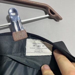 May include: Close-up of black trousers with a tag indicating adjustable waist sizes 39, 40, and 41 inches. The tag also provides instructions for adjusting the buckle. The trousers are hanging on a brown hanger.