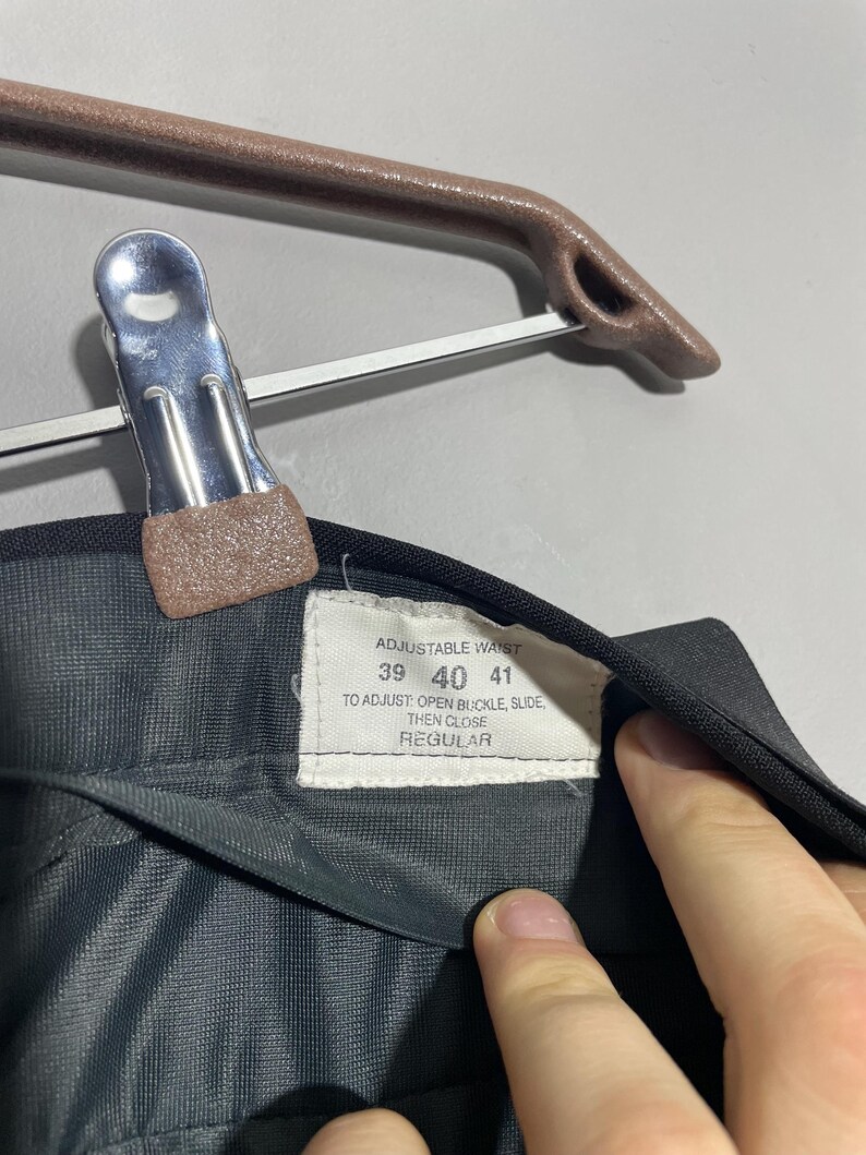 May include: Close-up of black trousers with a tag indicating adjustable waist sizes 39, 40, and 41 inches. The tag also provides instructions for adjusting the buckle. The trousers are hanging on a brown hanger.