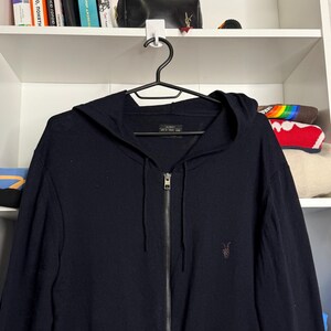 May include: A navy blue zip-up hoodie with a hood and a small brown logo on the chest. The hoodie is hanging on a black hanger against a white shelf background.