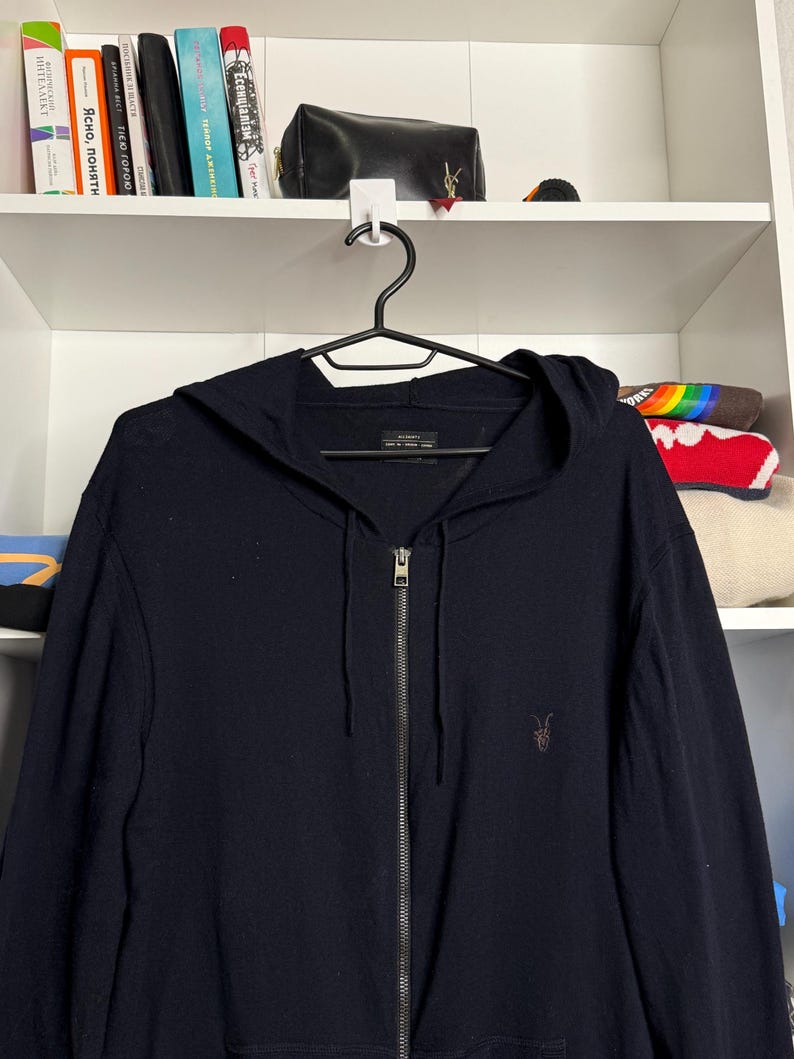 May include: A navy blue zip-up hoodie with a hood and a small brown logo on the chest. The hoodie is hanging on a black hanger against a white shelf background.
