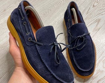 Puede incluir: Zapatos náuticos de ante azul marino con suelas color canela. Cordones y ojales de cuero. Diseño clásico de zapato náutico.