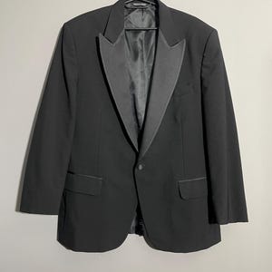 May include: A black tuxedo jacket with a satin lapel and single-button closure. The jacket has two front pockets and is hanging on a black hanger. The garment is a formal wear item.