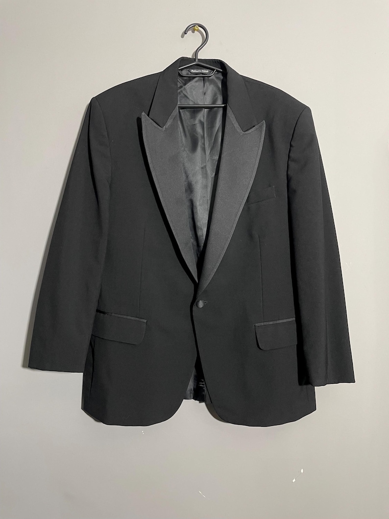 May include: A black tuxedo jacket with a satin lapel and single-button closure. The jacket has two front pockets and is hanging on a black hanger. The garment is a formal wear item.