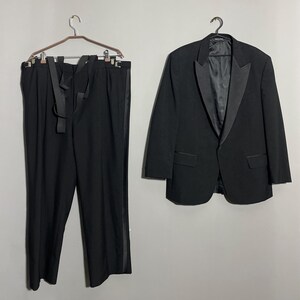 May include: A formal black tuxedo suit, including a jacket and trousers, hanging against a neutral background. The jacket has a satin lapel and the trousers have a satin stripe down the side. The suit is displayed on hangers.