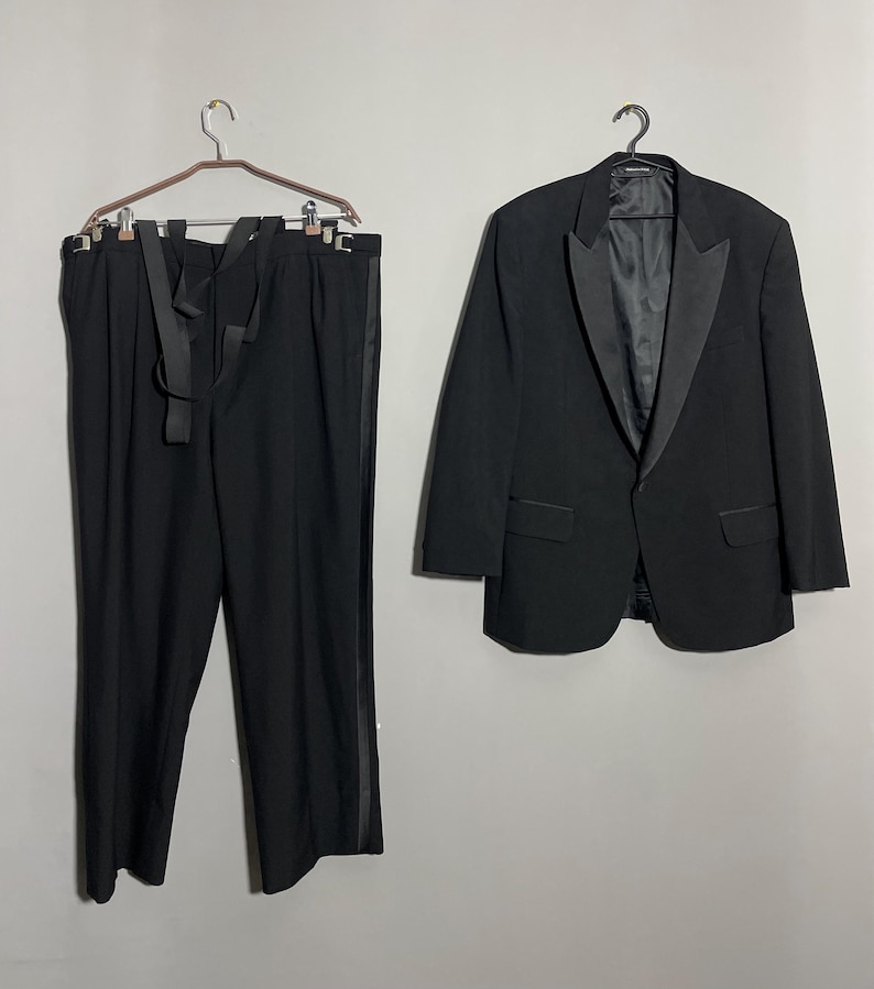May include: A formal black tuxedo suit, including a jacket and trousers, hanging against a neutral background. The jacket has a satin lapel and the trousers have a satin stripe down the side. The suit is displayed on hangers.