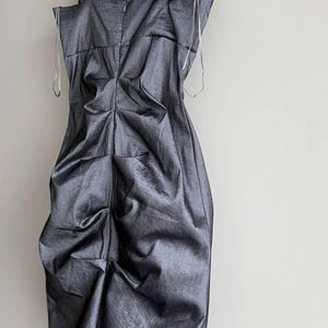 Puede incluir: Un vestido gris oscuro de un solo hombro con un dise&ntilde;o drapeado. El vestido cuelga de una percha de madera sobre un fondo blanco. La correa del hombro es negra y el vestido tiene un aspecto texturizado y met&aacute;lico.