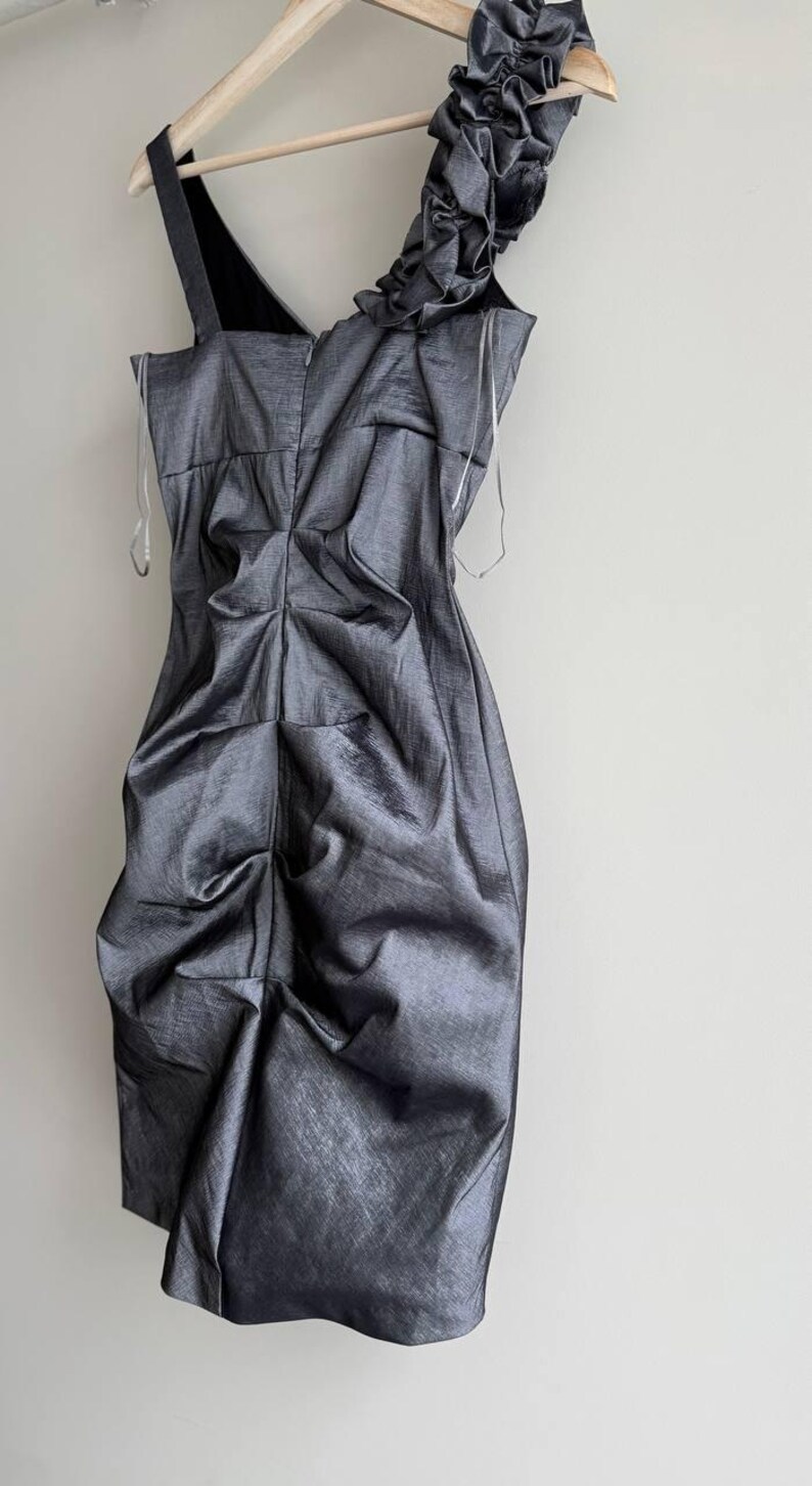 Puede incluir: Un vestido gris oscuro de un solo hombro con un dise&ntilde;o drapeado. El vestido cuelga de una percha de madera sobre un fondo blanco. La correa del hombro es negra y el vestido tiene un aspecto texturizado y met&aacute;lico.