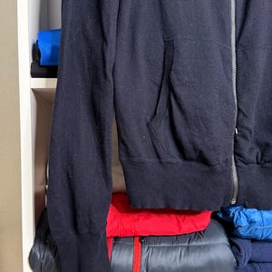 May include: A navy blue zip-up hoodie with a front pocket and a silver zipper. The hoodie is made of a soft material and is folded on a white shelf. Other folded clothing items in red, blue, and black are visible.