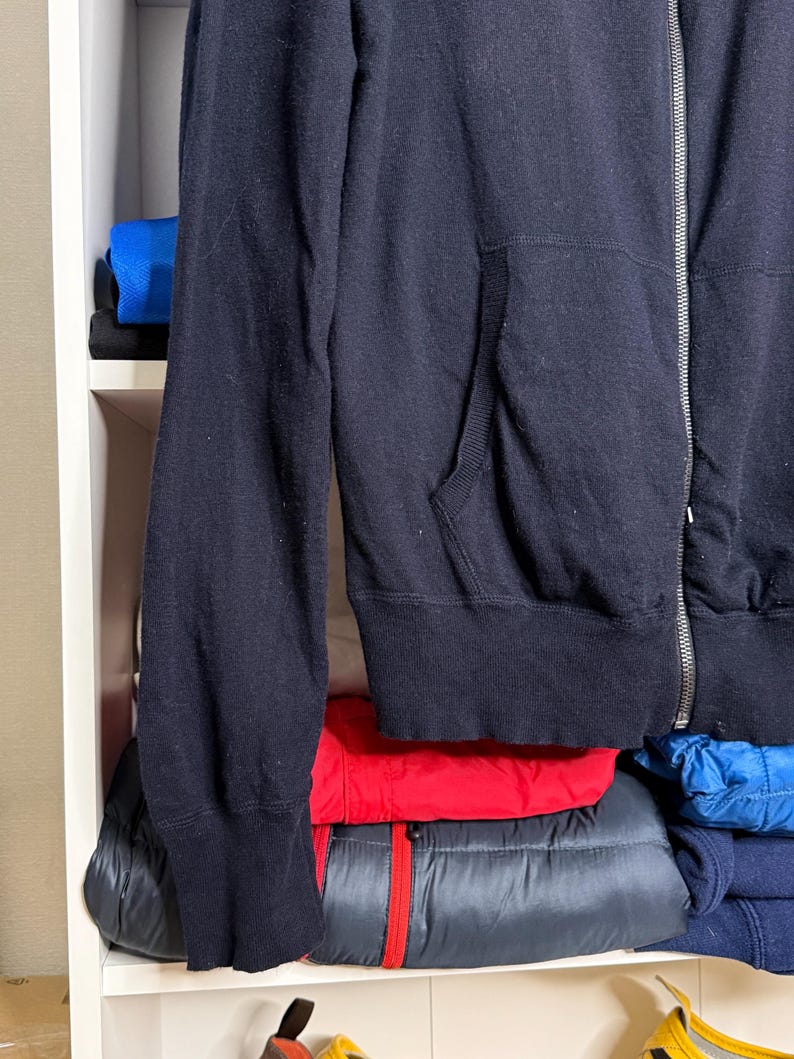 May include: A navy blue zip-up hoodie with a front pocket and a silver zipper. The hoodie is made of a soft material and is folded on a white shelf. Other folded clothing items in red, blue, and black are visible.