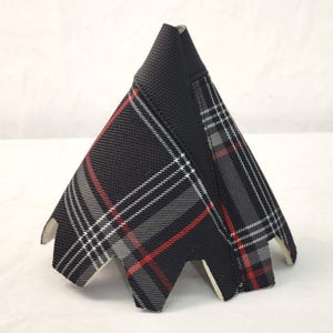 May include: A black and grey plaid fabric car gear stick gaiter with red accent lines. The gaiter has a triangular shape with a textured top and a cut-out base. The plaid pattern is a classic design.
