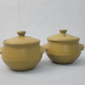 May include: Two small, round, tan ceramic pots with lids. Each pot has a handle on either side and a knob on the lid. The pots are on a white surface.