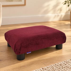 May include: A rectangular footstool upholstered in deep burgundy velvet fabric. The piece features four short, cylindrical black legs and a small fabric tag with a Union Jack flag design attached to the lower side edge of the cushion.