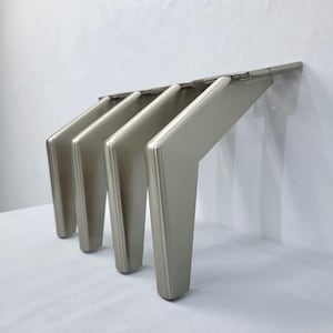May include: A set of four silver-coloured furniture legs. Each leg has a trapezoidal shape with a flat top and a slightly rounded bottom. The legs are made of metal and have a brushed finish. They are designed to be attached to a piece of furniture.