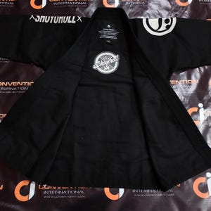 May include: Black martial arts gi with white logos and text. The gi has a collar and lapel, with the brand name "Shoyoroll" on a circular patch. The gi is spread open, showing the inside lining. The text "SNUTUKULLX" is visible on the shoulder.