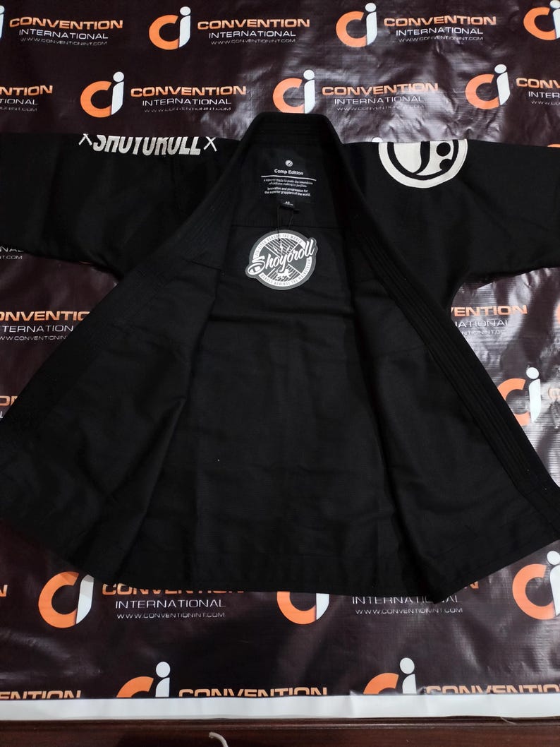 May include: Black martial arts gi with white logos and text. The gi has a collar and lapel, with the brand name "Shoyoroll" on a circular patch. The gi is spread open, showing the inside lining. The text "SNUTUKULLX" is visible on the shoulder.