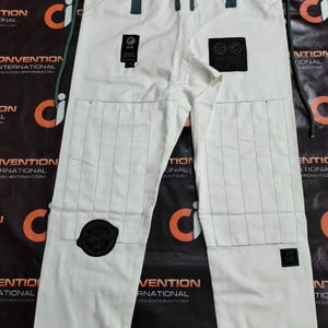 Op de afbeelding: Witte Jiu-Jitsu broek met zwarte stiksels en patches. De broek heeft een tailleband met trekkoord en versterkte knie&euml;n. Patches bevatten de tekst "SHOYOROLL JIU JITSU" en andere logo's. Ontworpen voor vechtsporttraining.