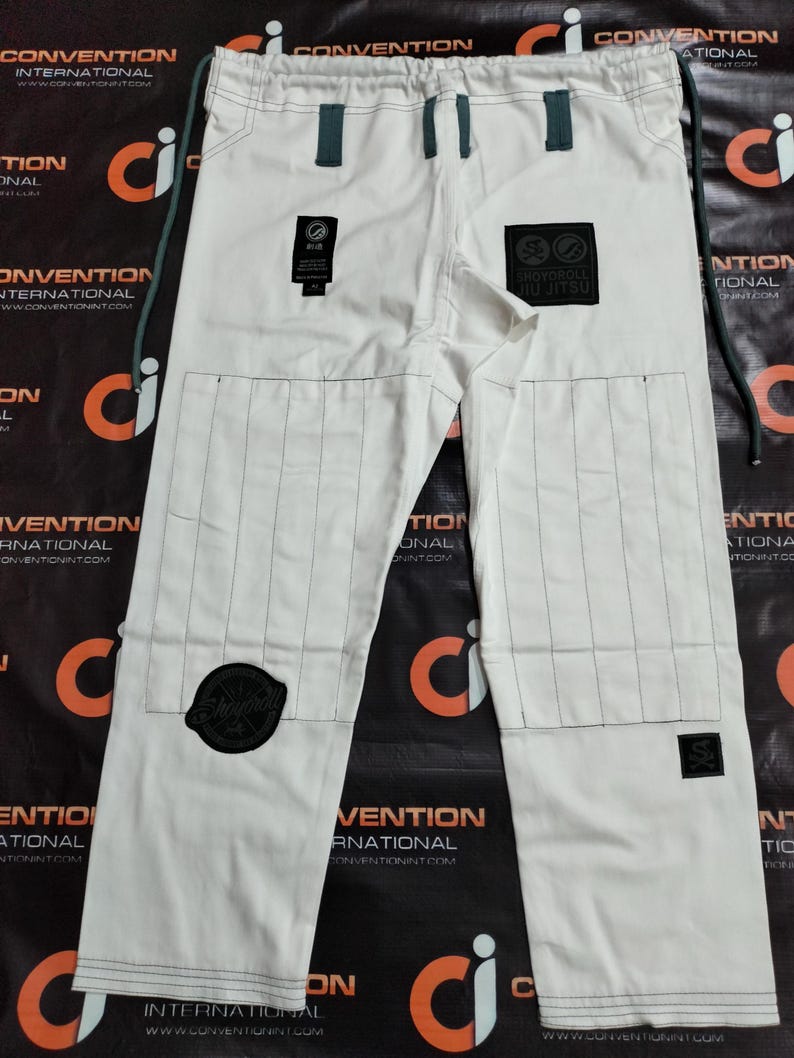 Op de afbeelding: Witte Jiu-Jitsu broek met zwarte stiksels en patches. De broek heeft een tailleband met trekkoord en versterkte knie&euml;n. Patches bevatten de tekst "SHOYOROLL JIU JITSU" en andere logo's. Ontworpen voor vechtsporttraining.