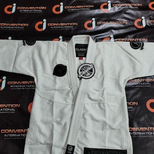 Op de afbeelding: Een witte Jiu-Jitsu gi met zwarte accenten, met het "Shoyoroll" logo en het "Classic" label. De gi heeft versterkte stiksels en een zwarte patch op de borst. De achtergrond heeft een herhalend logopatroon.