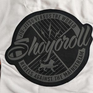 Op de afbeelding: Close-up van een wit Jiu-Jitsu-uniform met een grote, ronde zwarte patch. De patch bevat de woorden "Shoyoroll", "Jiu-Jitsu Versus The World" en "Rebels Against The Mainstream". Een kleinere "REBEL" patch is zichtbaar.