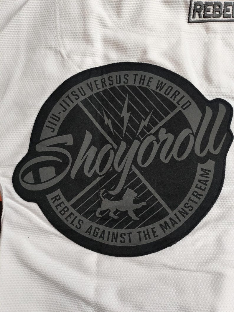 Op de afbeelding: Close-up van een wit Jiu-Jitsu-uniform met een grote, ronde zwarte patch. De patch bevat de woorden "Shoyoroll", "Jiu-Jitsu Versus The World" en "Rebels Against The Mainstream". Een kleinere "REBEL" patch is zichtbaar.