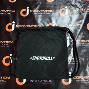 May include: Black drawstring bag with the white text "*SHOYOROLL*". The bag has black drawstrings and is set against a black background with orange and white logos. The bag is likely for carrying items.
