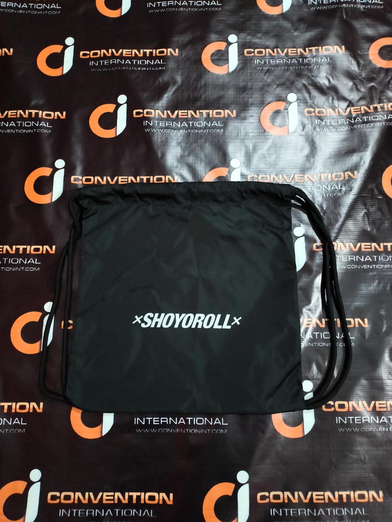 May include: Black drawstring bag with the white text "*SHOYOROLL*". The bag has black drawstrings and is set against a black background with orange and white logos. The bag is likely for carrying items.