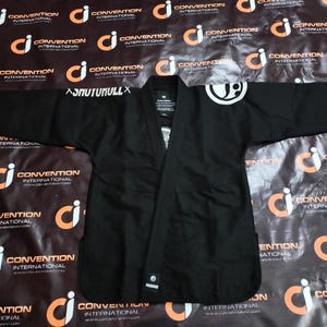 May include: Black Jiu-Jitsu gi with white logo on the shoulder and the text "ASNUTUKULLX" on the sleeve. The gi is laid flat, showcasing its design and construction. The background features a repeating pattern of a logo and the words "CONVENTION INTERNATIONAL".
