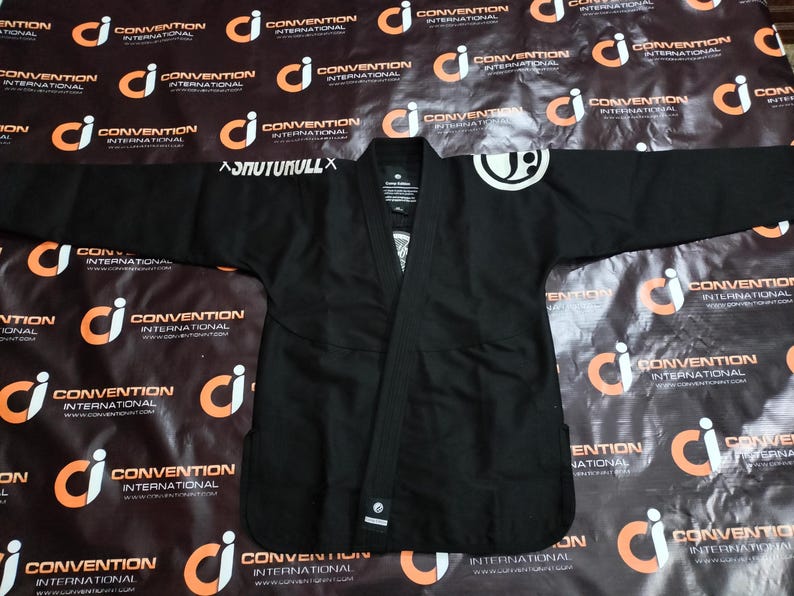 May include: Black Jiu-Jitsu gi with white logo on the shoulder and the text "ASNUTUKULLX" on the sleeve. The gi is laid flat, showcasing its design and construction. The background features a repeating pattern of a logo and the words "CONVENTION INTERNATIONAL".