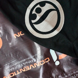 May include: Black fabric with a white and black circular emblem. The emblem features a stylized design within a circle. The word "NC" is visible in the image. The background includes a banner with the words "CONVENTION INTERNATIONAL".