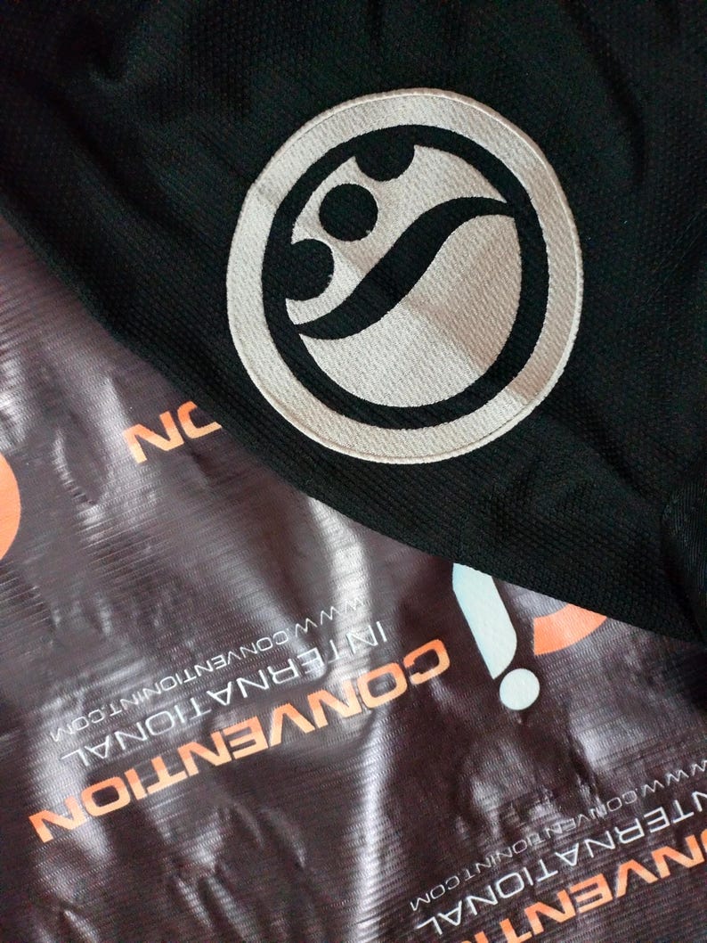 May include: Black fabric with a white and black circular emblem. The emblem features a stylized design within a circle. The word "NC" is visible in the image. The background includes a banner with the words "CONVENTION INTERNATIONAL".