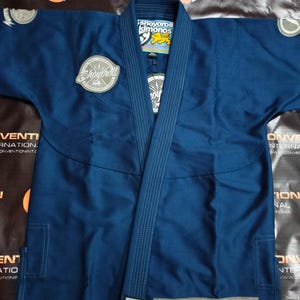 May include: A navy blue Shoyoroll Jiu-Jitsu gi. The gi features a jacket and trousers, with reinforced stitching and patches. The jacket has a lapel and is adorned with multiple Shoyoroll patches. The gi is designed for martial arts training and competition.
